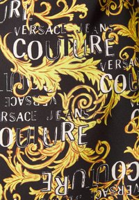 Tissu noir avec texte blanc "VERSACE JEANS COUTURE" répété et superposé de motifs floraux baroques ornés en jaune et orange.