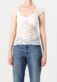 Haut en dentelle blanche avec de courtes manches gigot, présentant un motif floral et une encolure en V. Porté avec un jean en denim bleu.