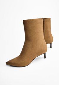 Paire de bottines en daim marron à bouts pointus, fermetures éclair latérales et talons fins et bas sur fond blanc.