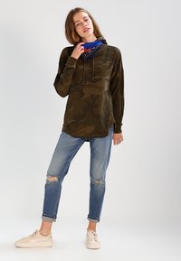 Camo-mønstret oversized hettegenser i mørkegrønn, kombinert med slitte lyseblå jeans. Modellen har på seg et blått bandana rundt halsen og beige joggesko.
