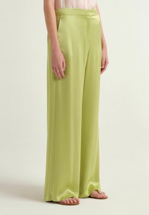 Donna che indossa pantaloni larghi a vita alta in raso verde lime con tasche laterali e sandali piatti marroni, in piedi contro uno sfondo chiaro uniforme.