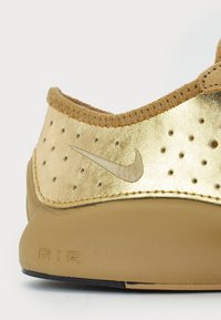 Kaošak zlatnog i bež tenisica sa perforacijama, Nike swoosh logom i teksturiranom đonom na kojem je otisnuto "AIR".