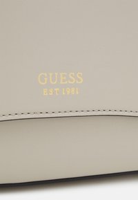 Borsa in ecopelle grigia con logo impresso "GUESS EST 1981" in oro. Texture liscia con cuciture scure a contrasto lungo i bordi.