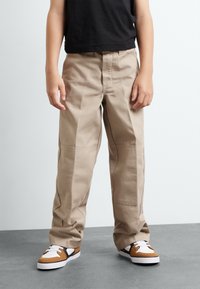 Pantalons en coton beige avec une coupe décontractée, dotés de coutures latérales, de jambes droites et de couture visible. Assortis avec des baskets marron et blanches.