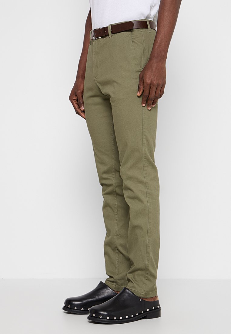 Olivgröna byxor med rak passform och ett brunt läderbälte. Tyget är slätt med en lätt lyster. Svarta träskor med silverdetaljer.