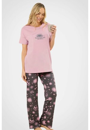 Femme souriante portant un t-shirt rose "WEEKEND DREAMING" et un pantalon de pyjama foncé avec des motifs de soleil et de lune roses, tenant une tasse grise, debout pieds nus.