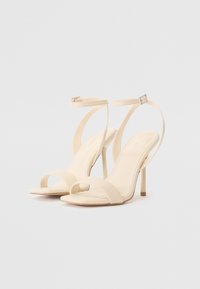 Beige høyhælte sandaler med stropper, med et slankt design, som har en tynn ankelrem, åpen tå og glatt tekstur.