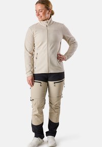 Beige fleecejacka med hög krage, fickor med dragkedja; beige och svarta zip-off byxor med dragkedjor och logodetaljer; vita sneakers.