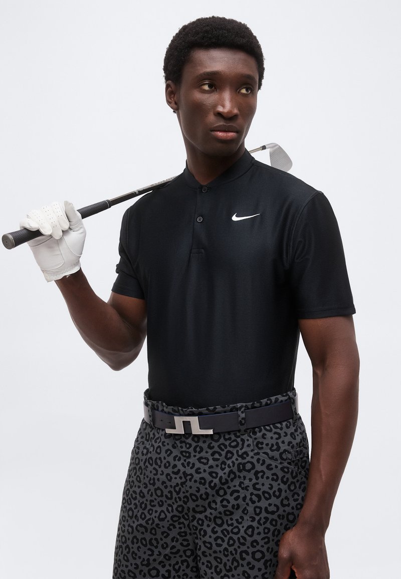 Homme portant un polo Nike noir, un pantalon à imprimé léopard et un gant de golf blanc, tenant un club de golf reposant sur l’épaule sur fond blanc.
