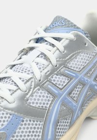 Gros plan d'une chaussure de course Asics blanche et bleu clair avec un tissu en mesh et des lacets blancs noués en motif croisé.