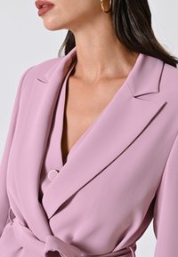 Helles rosa Blazer mit Schalkragen, Wickeldesign und einem kreisförmigen silbernen Akzent am Verschluss. Glattes, strukturiertes Gewebe mit einer figurbetonten Silhouette.