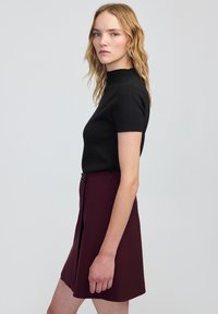 Zwarte top met korte mouwen en hoge hals, gecombineerd met een bordeauxrode A-lijn rok. De outfit heeft een zachte textuur en knoopdetails aan de voorkant.