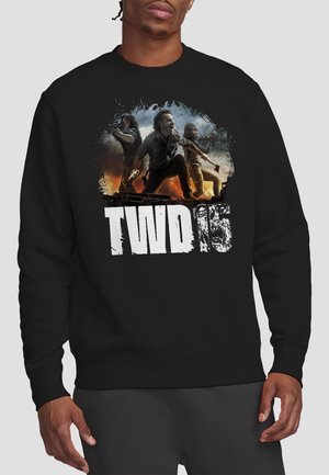 Three armed characters in action poses with "TWD 15" text on a black sweatshirt worn by a person.