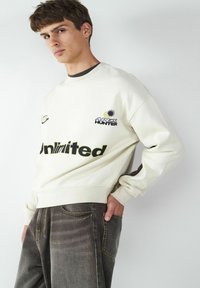 Kremowa bluza z czarnym napisem "Unlimited" oraz logo "APEX HUNTER", z okrągłym dekoltem i ściągaczami. Noszona z szarymi dżinsami.