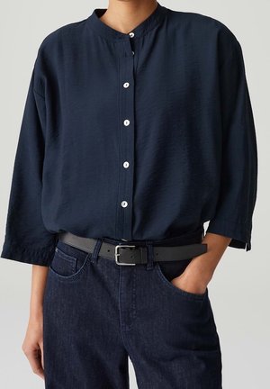 Personne portant une chemise bleu marine boutonnée aux manches trois-quarts, rentrée dans un jean en jean foncé avec une ceinture noire. Une main dans la poche.