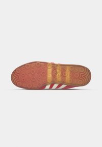 Röd sportskosula med cirkulära mönster; har tre vita ränder, texturerad gummi för grepp och en präglad logotyp i mitten.