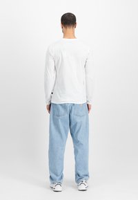 Langermet hvit t-skjorte med rund hals, kombinert med lyseblå løstsittende jeans og hvite sneakers, sett fra baksiden.