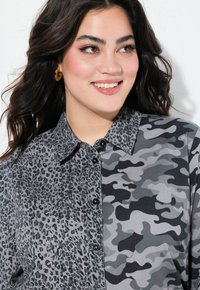 Ung kvinde, der smiler, iført en skjorte med knapper i blandede grå leopard- og camouflage-mønstre samt gyldne hoops.