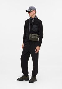 Μαύρη μπλούζα με μακριά μανίκια και τσέπη στο στήθος, συνδυασμένη με χαλαρές μαύρες παντελόνες. Γκρι καπέλο, μαύρο μαντήλι και ελαιόχρωμη τσάντα χιαστί. Χοντρές μαύρες παπούτσια.