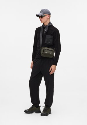 Schwarzes langärmliges Oberteil mit einer Brusttasche, kombiniert mit lockeren schwarzen Hosen. Graue Mütze, schwarzes Bandana und olivgrüne Umhängetasche. Bulky schwarze Schuhe.