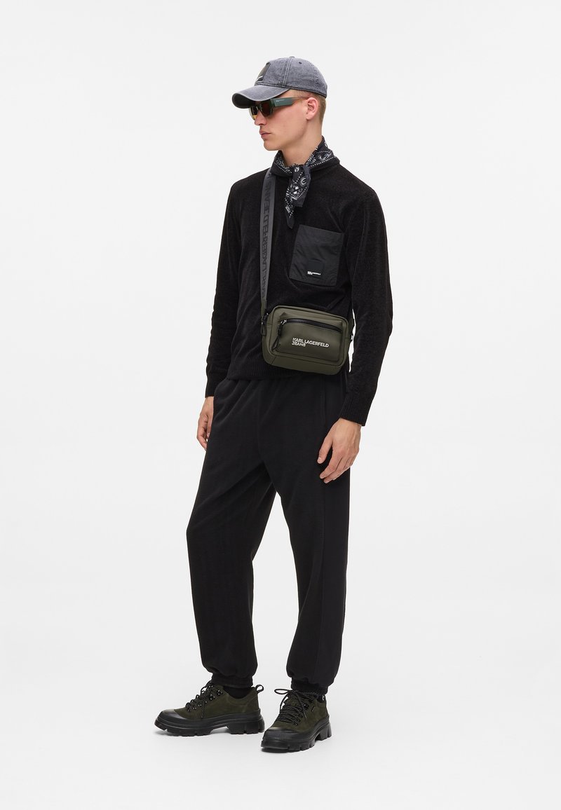 Μαύρη μπλούζα με μακριά μανίκια και τσέπη στο στήθος, συνδυασμένη με χαλαρές μαύρες παντελόνες. Γκρι καπέλο, μαύρο μαντήλι και ελαιόχρωμη τσάντα χιαστί. Χοντρές μαύρες παπούτσια.