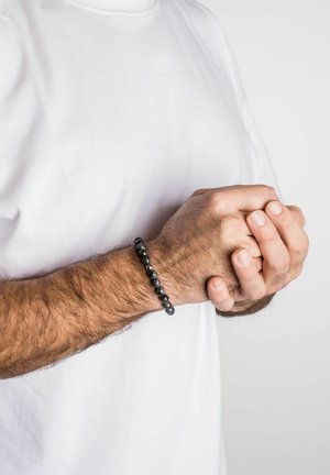 Schwarzes Perlenarmband aus polierten Steinen, in runder Form, getragen an einem Handgelenk mit heller Hautfarbe und hellfarbener Kleidung.