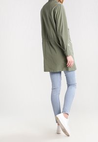 Giacca verde oliva a maniche lunghe con vita arricciata, indossata sopra jeans attillati azzurri e sneakers bianche, con collo con bottoni e vestibilità comoda.