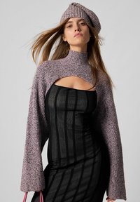 Kvinde iført en sort gennemsigtig stribet kjole med en udskæring over brystet, en lyserød strikket cropped sweater og en matchende strikket beret.