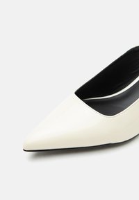 Λευκό μυτερό γυναικείο παπούτσι με τακούνι και μαύρη εσωτερική επένδυση σε λευκό φόντο.