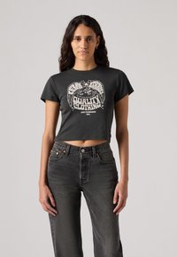 Gekürztes schwarzes Baumwoll-T-Shirt mit einem grafischen Druck, auf dem "QUALITÄTSKLEIDUNG" steht, kombiniert mit hochgeschnittenen dunklen Jeans.