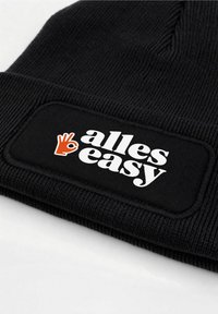 Black beanie hat with ribbed texture, featuring a rectangular white patch that reads "alles easy" and an orange hand symbol.