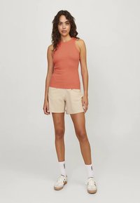 Lightweight, sleeveless, rust-colored tank top paired with beige shorts. Model wears white socks and cream sneakers, standing against a plain background.