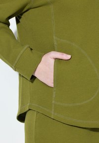 Hnědozelený textured pletený top s boční kapsou, zaobleným lemem a kontrastním prošitím podél švů.