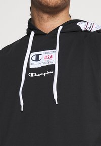 Black hoodie made of soft fabric featuring white drawstrings. Displays prominent "Champion" logo and layered brand labels on the chest.