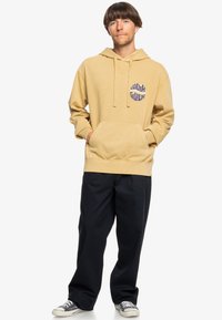 Beige cotton hoodie with front pocket, drawstring hood, and circular "Quiksilver" logo in blue. Paired with black pants and sneakers.