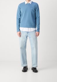 Hellblaues Sweatshirt mit V-Ausschnitt, über einem weißen Hemd mit Kragen getragen, kombiniert mit hellblauen Jeans und schwarzen Schuhen. Schlichtes und lässiges Design.