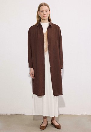 Nő áll egy sima fal előtt, hosszú barna gombos ingben, fehér ruhában, türkiz fülbevalóval és barna hegyes orrú balerinacipőben.