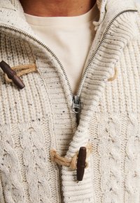 Cardigan in maglia a trecce color avorio con colletto alto, dotato di cerniera a tutta lunghezza e chiusure a toggle in legno, realizzato in tessuto di maglia testurizzato.