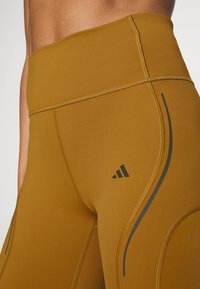 Leggings de tiro alto en color beige, elaborados con una tela elástica que presenta detalles en negro y costuras. Cuentan con un diseño de líneas curvas y un logo de adidas.