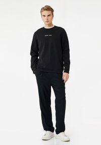 Sudadera negra con "Liberté Totale" bordado en blanco, combinado con pantalones negros y zapatillas blancas. Diseño simple y casual.