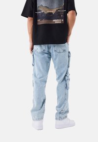 Jeans cargo de color azul claro con múltiples bolsillos laterales, que se estrechan en los tobillos, combinados con una camiseta gráfica negra y zapatillas blancas.