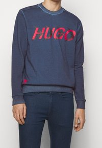 Felpa blu navy con polsini e orlo a costine, con un audace logo "HUGO" rosso decorato con motivi fogliari. Materiale morbido e scollatura a girocollo.