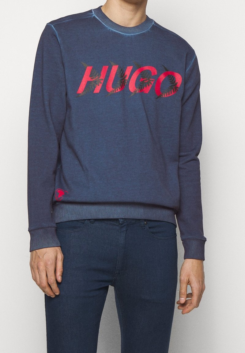 Felpa blu navy con polsini e orlo a costine, con un audace logo "HUGO" rosso decorato con motivi fogliari. Materiale morbido e scollatura a girocollo.