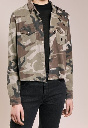 Osoba v krátké maskáčové bundě s předními kapsami a černými flitry přes černé tričko a tmavé džíny, zobrazena od krku po boky.