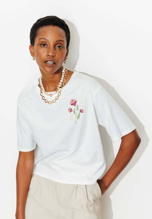 Femme aux cheveux courts portant un t-shirt blanc avec une broderie florale rose et violette, un pantalon beige, et des colliers superposés en or et perles.