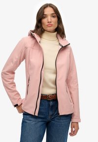 Chaqueta rosa suave hecha de tela ligera y lisa con capucha, cierre frontal con cremallera, bolsillos laterales y detalles en negro; llevada sobre un suéter de cuello alto beige.