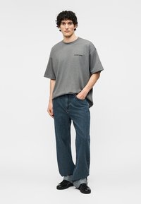 Joven con cabello rizado, vestido con una camiseta gris suelta, jeans azules de pierna ancha y zapatos negros, de pie contra un fondo liso.