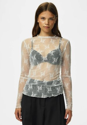 Ung kvinde med langt brunt hår iført en gennemsigtig hvid blomstret blonder-top over en grå bh og sorte bukser, stående mod en ensfarvet baggrund.