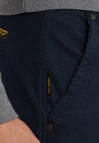 Pantalons en velours côtelé bleu marine avec des poches structurées et une étiquette de logo. Le tissu présente un motif côtelé texturé, avec un détail de bouton métallique.