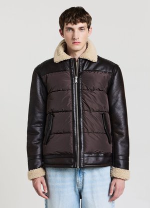 Giovane uomo che indossa una giacca imbottita marrone scuro con maniche in pelle nera e colletto e polsini in shearling beige, abbinata a jeans azzurri.